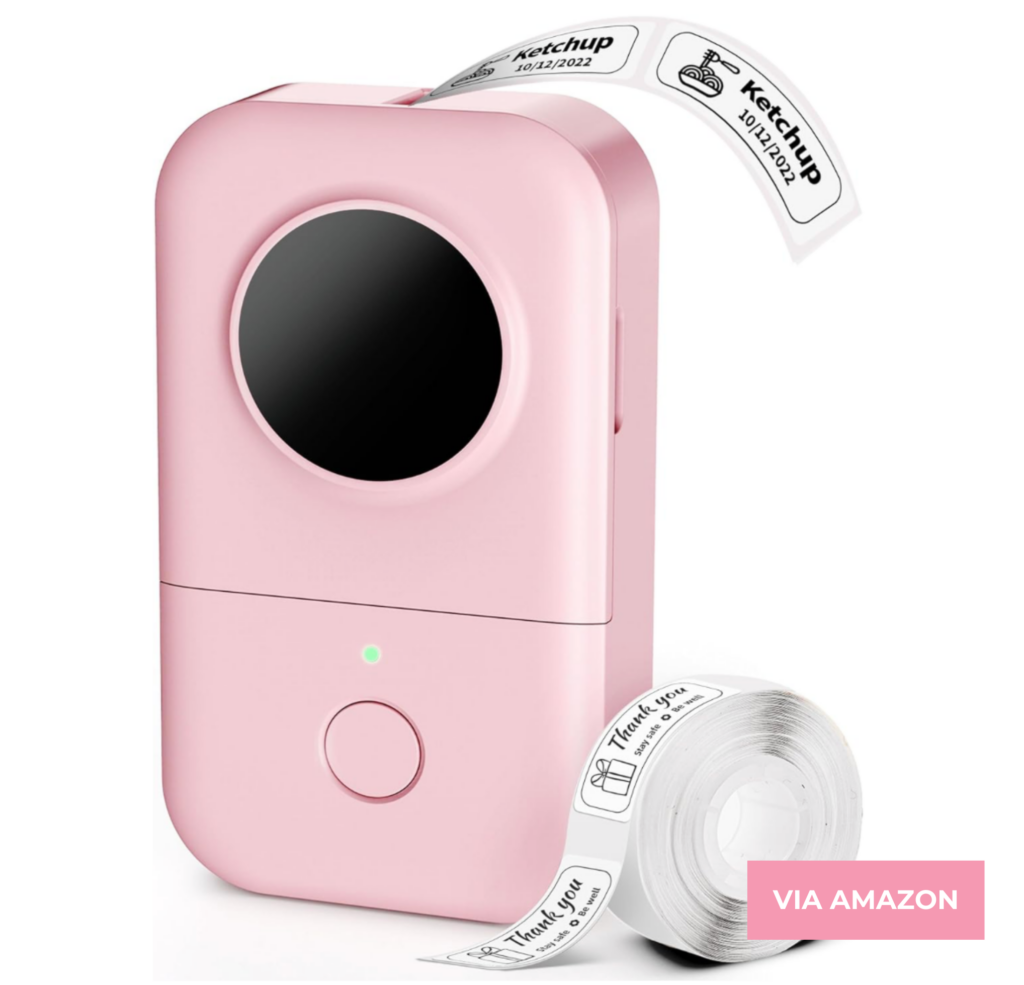 A pink portable label printer with a circular screen. Labels with text, including "Ketchup," are being printed. A roll of labels sits next to the printer. Text on the image says "VIA AMAZON.