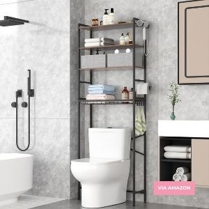 Metal bathroom shelf unit stands over a white toilet, holding towels, toiletries, and decorative items in a modern, tiled bathroom. A "VIA AMAZON" label appears in the image.